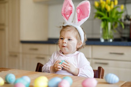 Foto de Linda niña pequeña con orejas de conejo de Pascua jugando con huevos pastel de colores. Niño feliz desempacando regalos. Adorable niño sonriente sano en ropa rosa disfrutando de unas vacaciones en familia, - Imagen libre de derechos