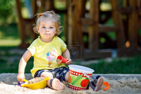 Foto de Happy toddler girl playing in sand on outdoor playground. Baby having fun on sunny warm summer sunny day. Active child with sand toys and in colorful fashion clothes - Imagen libre de derechos