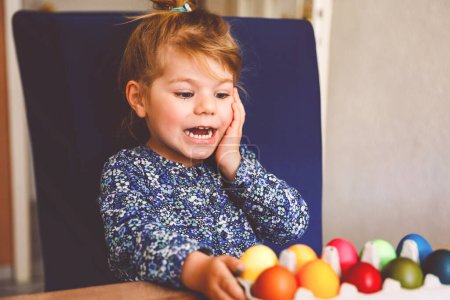 Photo for Excited little toddler girl coloring eggs for Easter. Child looking surprised at colored egg hoarding and celebrating catholic and christian holiday with family. Cute kid helping to color, indoors - Royalty Free Image