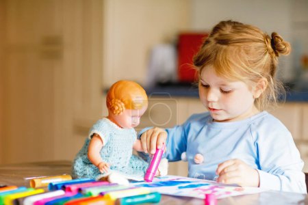 Photo for Little toddler girl painting with felt pens during pandemic coronavirus quarantine disease. Happy creative child with old vintage doll, homeschooling and home daycare with parents - Royalty Free Image
