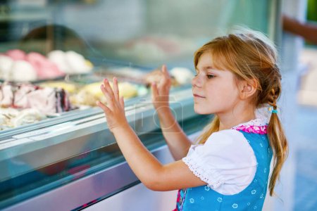 Téléchargez les photos : Cute little preschool girl choosing and buying ice cream in outdoor stand cafe. Happy smiling child looking at different sorts of icecream. Sweet summer dessert. - en image libre de droit