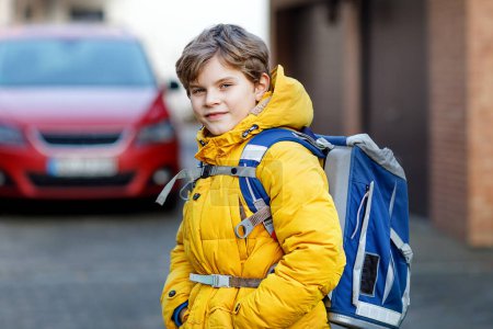 Téléchargez les photos : Petit écolier du primaire marchant jusqu'à l'école. Portrait d'enfant heureux dans la rue avec la circulation. Étudiant avec veste jaune et sac à dos en vêtements d'hiver colorés - en image libre de droit