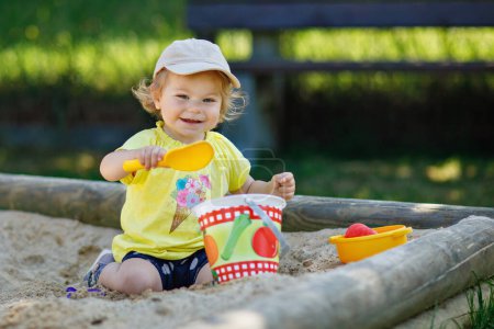 Téléchargez les photos : Bonne petite fille jouant dans le sable sur l'aire de jeux extérieure. Bébé s'amuser par une chaude journée ensoleillée d'été. Enfant actif avec des jouets de sable et des vêtements de mode colorés - en image libre de droit