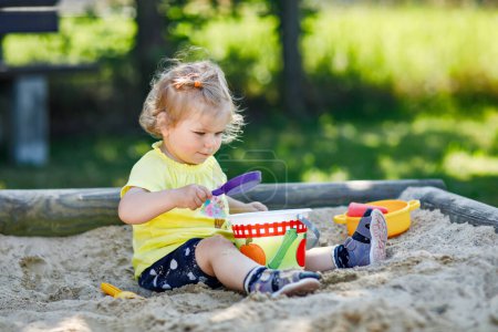 Foto de Happy toddler girl playing in sand on outdoor playground. Baby having fun on sunny warm summer sunny day. Active child with sand toys and in colorful fashion clothes - Imagen libre de derechos