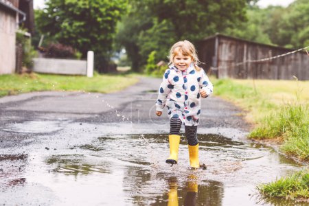 Foto de Niña pequeña con botas de lluvia amarillas, corriendo y caminando durante el aguanieve en el día nublado lluvioso. Lindo niño feliz en ropa colorida saltando en el charco, salpicaduras de agua, actividad al aire libre. - Imagen libre de derechos
