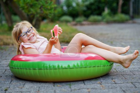 Téléchargez les photos : Cute little girl with glasses eating watermelon on inflatable ring in summertime. Happy smiling preschool child having fun. Healthy summer food and snacks for kids - en image libre de droit