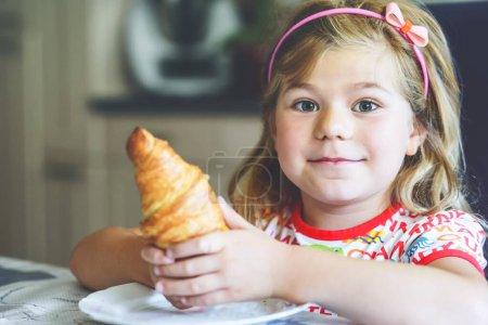 Téléchargez les photos : Enfant souriant au petit déjeuner. Nourriture et enfants heureux. La fille mange un croissant. Jolie fille préscolaire ayant un repas sain - en image libre de droit