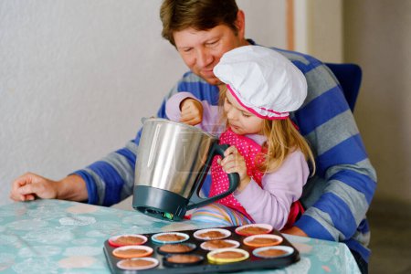 Téléchargez les photos : Mignon petit tout-petit fille et père cuisson maison muffins au chocolat cupcakes à la maison à l'intérieur. Joyeux enfant d'âge préscolaire et papa ensemble dans la cuisine domestique - en image libre de droit