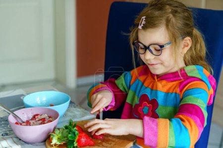 Téléchargez les photos : Littl maternelle apprendre à utiliser un couteau coupe fraise fabrication de salade de fruits pour la bouillie pour le petit déjeuner dans la cuisine. Aliments sains pour enfants - en image libre de droit