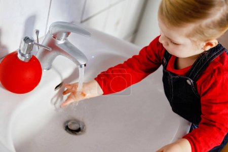 Foto de Linda niña pequeña lavándose las manos con agua y jabón en el baño. Adorable niño aprendiendo a limpiar partes del cuerpo. Rutina de higiene. Feliz niño sano en casa o guardería - Imagen libre de derechos