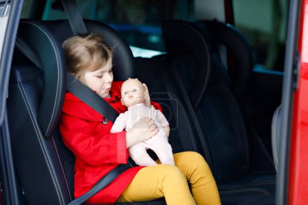 Photo for Toddler girl sitting in car seat, holding favourite doll toy and looking out of the window on nature and traffic. Little kid traveling by car. Child safety on the road. Family trip and vacations. - Royalty Free Image