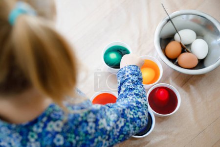 Photo for Closeup of little toddler girl coloring eggs for Easter. Close-up of child looking surprised at colorful colored eggs, celebrating holiday with family. From above, unrecognized face - Royalty Free Image
