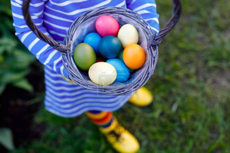 Photo for Close-up of of hands of toddler holding basket with colored eggs. Child having fun with traditional Easter eggs hunt, outdoors. Celebration of christian holiday. - Royalty Free Image