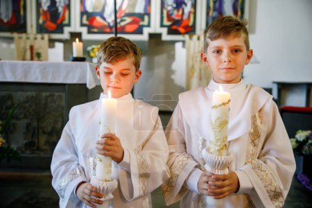 Photo for Two little kids boys receiving his first holy communion. Happy children holding Christening candle. Tradition in catholic curch. Kids in a church near altar. Siblings, brothers in white gowns - Royalty Free Image