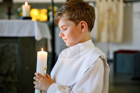 Petit garçon recevant sa première sainte communion. Joyeux enfant tenant une bougie du baptême. Tradition en curch catholique. Enfant en robe traditionnelle blanche dans une église près de l'autel

