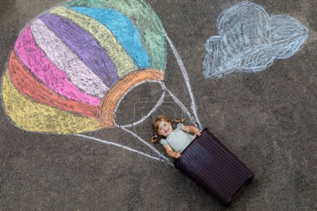 Téléchargez les photos : Joyeux petit enfant volant en montgolfière peint avec des craies colorées aux couleurs arc-en-ciel sur le sol ou de l'asphalte en été. Mignon enfant qui s'amuse. loisirs créatifs pour les enfants - en image libre de droit