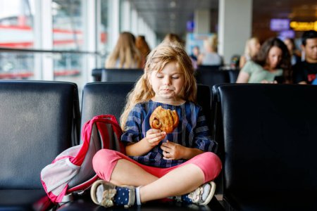 Téléchargez les photos : Petite fille à l'aéroport attendant l'embarquement à la grande fenêtre. Un mignon gosse qui mange un chignon pour une collation. Au plaisir de partir pour des vacances d'été en famille. - en image libre de droit