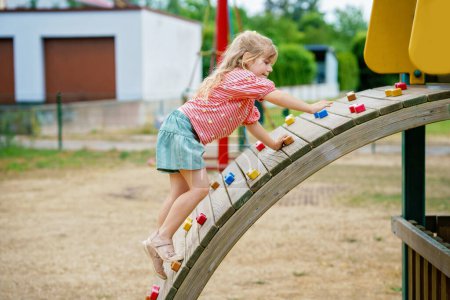 Téléchargez les photos : Petite fille grimpant sur l'aire de jeux le jour de l'été. Happy brave enfant d'âge préscolaire appréciant le sport et l'activité des enfants - en image libre de droit