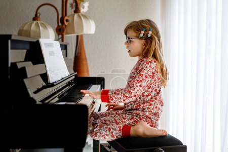 Niña feliz tocando el piano en la sala de estar. Lindo niño preescolar con gafas para los ojos que se divierten con aprender a tocar el instrumento de música
