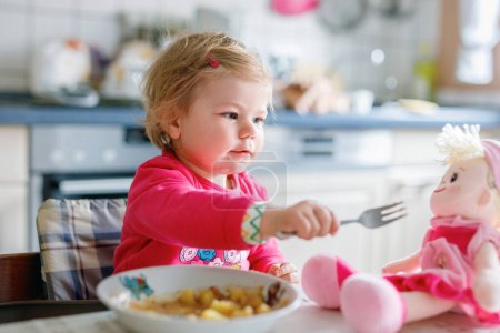 Foto de Adorable niña comiendo verduras de tenedor y pasta. alimento, niño, alimentación y desarrollo. Lindo niño, hija con cuchara sentada en silla alta y aprender a comer por sí mismo - Imagen libre de derechos