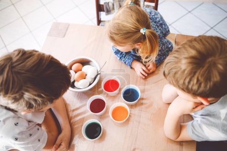 Téléchargez les photos : Petite fille tout-petit excitée et deux enfants garçons colorant des œufs pour Pâques. Trois enfants, frères et sœurs regardant surpris par des œufs colorés, célébrant des vacances en famille. D'en haut, visages méconnus. - en image libre de droit