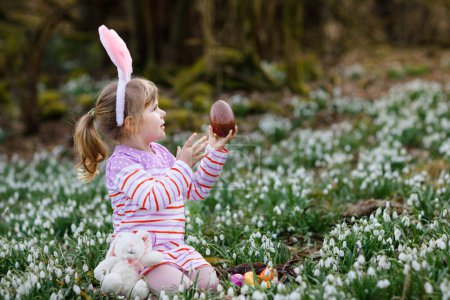 Foto de Niña con orejas de conejo de Pascua haciendo caza de huevos en el bosque de primavera en un día soleado, al aire libre. Lindo niño feliz con un montón de flores de la gota de nieve, enorme huevo de chocolate y huevos de colores - Imagen libre de derechos