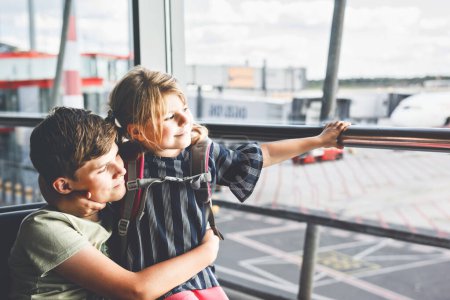Téléchargez les photos : Petite fille et écolier à l'aéroport attendant l'embarquement à la grande fenêtre. Deux enfants se tiennent à la fenêtre sur fond d'avions. Enfants heureux, frères et sœurs partent pour des vacances d'été en famille. - en image libre de droit