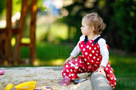 Téléchargez les photos : Petite fille mignonne jouant dans le sable sur une aire de jeux extérieure. Beau bébé en chewing-gum rouge qui s'amuse par une chaude journée d'été ensoleillée. Enfant avec des jouets de sable colorés. Bébé actif sain à l'extérieur joue à des jeux - en image libre de droit