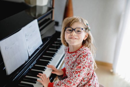 Foto de Little happy girl playing piano in living room. Cute preschool child with eye glasses having fun with learning to play music instrument - Imagen libre de derechos