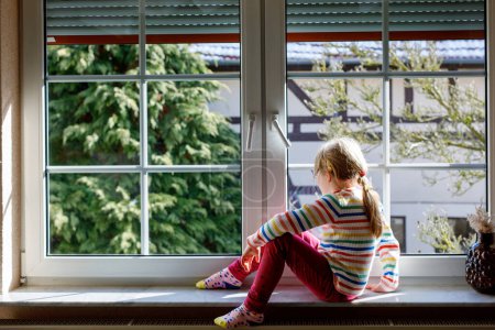 Foto de Adorable niñita sentada junto a la ventana. Niño preescolar feliz mirando hacia fuera y disfrutando de un día soleado - Imagen libre de derechos