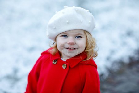 Téléchargez les photos : Portrait d'hiver en plein air de petite fille tout-petit mignonne en manteau rouge et barret chapeau de mode blanc. Bébé enfant heureux et en bonne santé marchant dans le parc par temps froid avec neige et chutes de neige. Des vêtements élégants pour les enfants - en image libre de droit