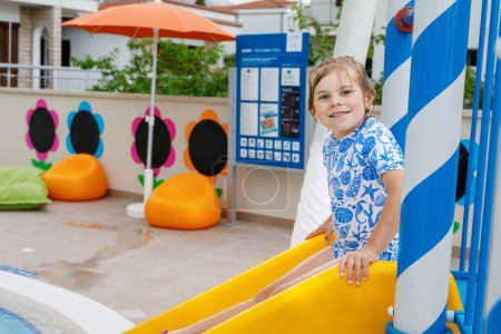 Photo for Little preschool girl sliding on a children slide in outdoor swimming pool in hotel resort. Child learning to swim in outdoor pool, splashing with water, laughing and having fun. Family vacations - Royalty Free Image