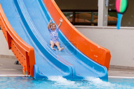 Téléchargez les photos : Petite fille d'âge préscolaire glissant sur une glissière d'enfants dans la piscine extérieure dans la station hôtelière. Enfant apprenant à nager dans la piscine extérieure, éclaboussant d'eau, riant et s'amusant. Vacances en famille - en image libre de droit