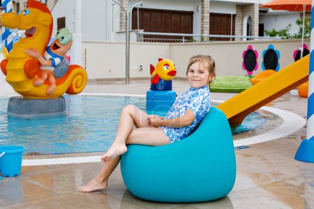 Foto de Niña preescolar sentada junto a la piscina al aire libre en el complejo hotelero. Niños aprendiendo a nadar en la piscina al aire libre, salpicando con agua, disfrutando, riendo y divirtiéndose. Vacaciones familiares - Imagen libre de derechos
