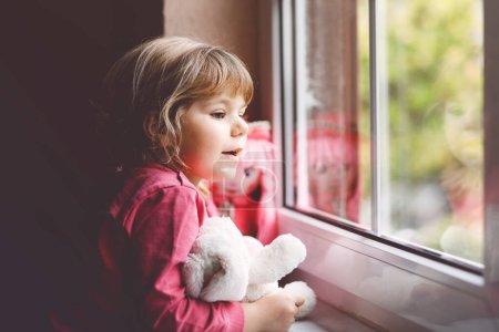 Photo for Cute toddler girl sitting by window and looking out on rainy day. Dreaming child with doll and soft toy feeling happy. Self isolation concept during corona virus pandemic time. Lonely kid - Royalty Free Image