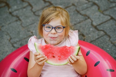 Foto de Cute little girl with glasses eating watermelon on inflatable ring in summertime. Happy smiling preschool child having fun. Healthy summer food and snacks for kids - Imagen libre de derechos