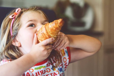 Photo for Smiling child at breakfast. Food and happy kids. The girl is eating a croissant. Cute preschool girl having healthy meal - Royalty Free Image