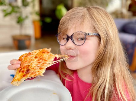 Téléchargez les photos : Petite fille d'âge préscolaire mangeant un morceau de pizza. Happy Child mordant pizza au fromage - en image libre de droit