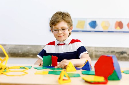 Foto de Niño pequeño con gafas jugando con elementos plásticos coloridos kit en la escuela o guardería preescolar. Feliz niño construyendo y creando figuras geométricas, aprendiendo matemáticas y geometría - Imagen libre de derechos