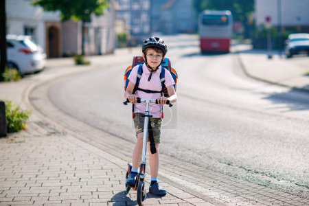 Photo for Active school kid boy with glasses in safety helmet riding with his scooter in the city with backpack on sunny day. Happy child biking on way to school. Safe way for kids outdoors to school - Royalty Free Image