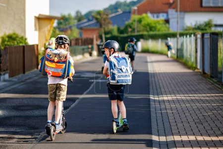 Téléchargez les photos : Deux garçons de l'école en casque de sécurité équitation avec scooter dans la ville avec sac à dos sur la journée ensoleillée. Enfants heureux en vêtements colorés faisant du vélo sur le chemin de l'école. Moyens sûrs pour les enfants à l'extérieur de l'école. - en image libre de droit