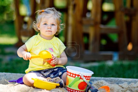 Foto de Feliz niña jugando en la arena en el patio al aire libre. Bebé divirtiéndose en el soleado verano cálido día soleado. Niño activo con juguetes de arena y en ropa de moda colorida - Imagen libre de derechos