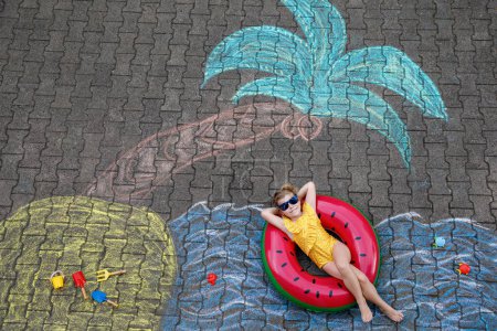 Téléchargez les photos : Happy little preschool girl in swimsuit on inflatable ring with sea, sand, palm painted with colorful chalks on asphalt. Cute child with having fun with chalk picture. Summer, vacations, summertime. - en image libre de droit