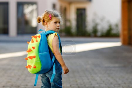 Photo for Cute little adorable toddler girl on her first day going to playschool. Healthy upset sad baby walking to nursery school. Fear of kindergarten. Unhappy child with backpack on city street, outdoors - Royalty Free Image