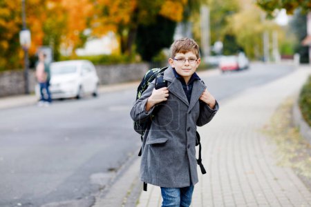 Photo for Happy kid boy with glasses and backpack or satchel. Schoolkid in stylish fashon coan on the way to middle or high school on cold autumn day. Healthy child outdoors on the street, on rainy day - Royalty Free Image