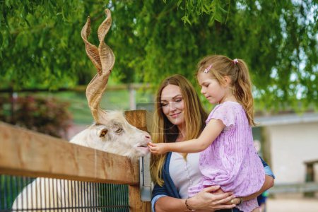 Photo for Little preschool girl and woman feeding goat. Happy excited child and mother feeds animals a wildlife park. Family leisure and activity for vacations or weekend. - Royalty Free Image