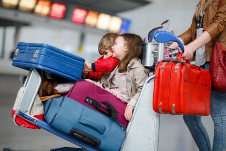 Foto de Dos niños pequeños, niño y niña, hermanos y madre en el aeropuerto. Niños, familia viajando, yendo de vacaciones en avión y esperando en el carro con maletas empujando por la mujer en la terminal para el vuelo - Imagen libre de derechos
