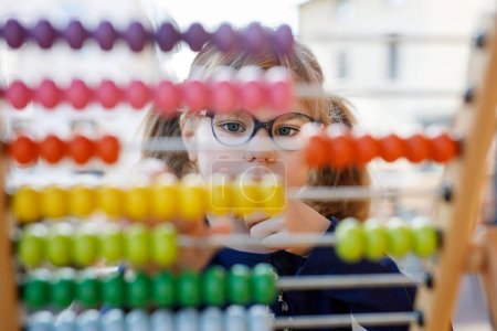 Little preschool girl playing with educational wooden rainbow toy counter abacus. Healthy happy child with glasses learning to count and colors, indoors on sunny day