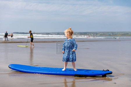Foto de Little cute toddler girl at the Ballybunion surfer beach, having fun on surfboard for the first time, west coast of Ireland. Happy child enjoying Irish summer with family - Imagen libre de derechos