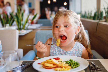 Foto de Adorable niña desayunando en el restaurante del resort. Feliz niño en edad preescolar comiendo alimentos saludables, verduras y huevos en la mañana - Imagen libre de derechos
